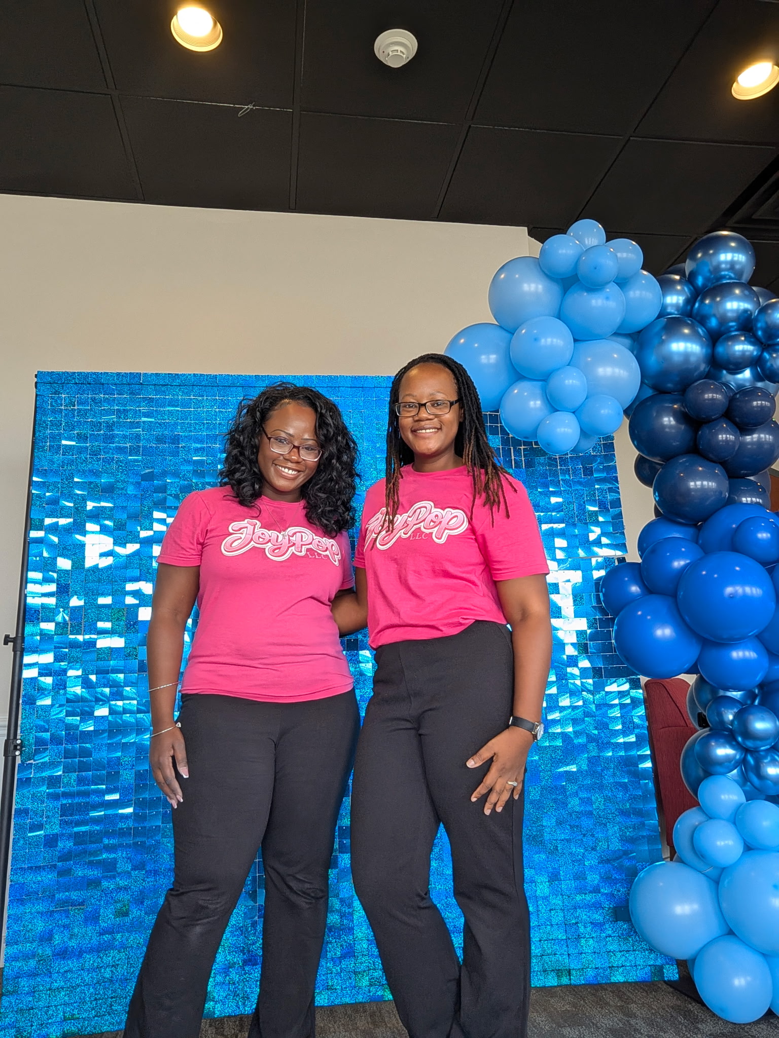The JoyPop team in pink shirts in front of a blue shimmer wall and balloon garland